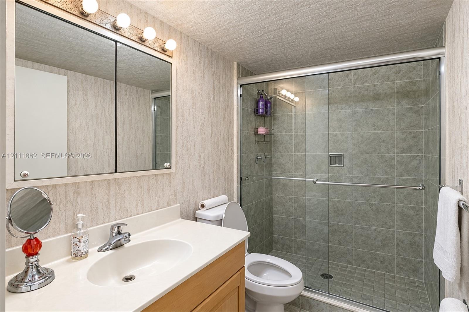Bathroom featuring a wood-finish vanity with a solid-surface countertop, an integrated sink, a mirrored medicine cabinet with integrated lighting, and a walk-in shower with a glass sliding door and tiled surround