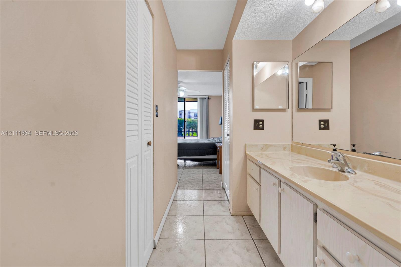 Bathroom featuring a long vanity with an integrated basin, light-toned cabinetry, a full-width mirror, tile flooring, and louvered closet doors