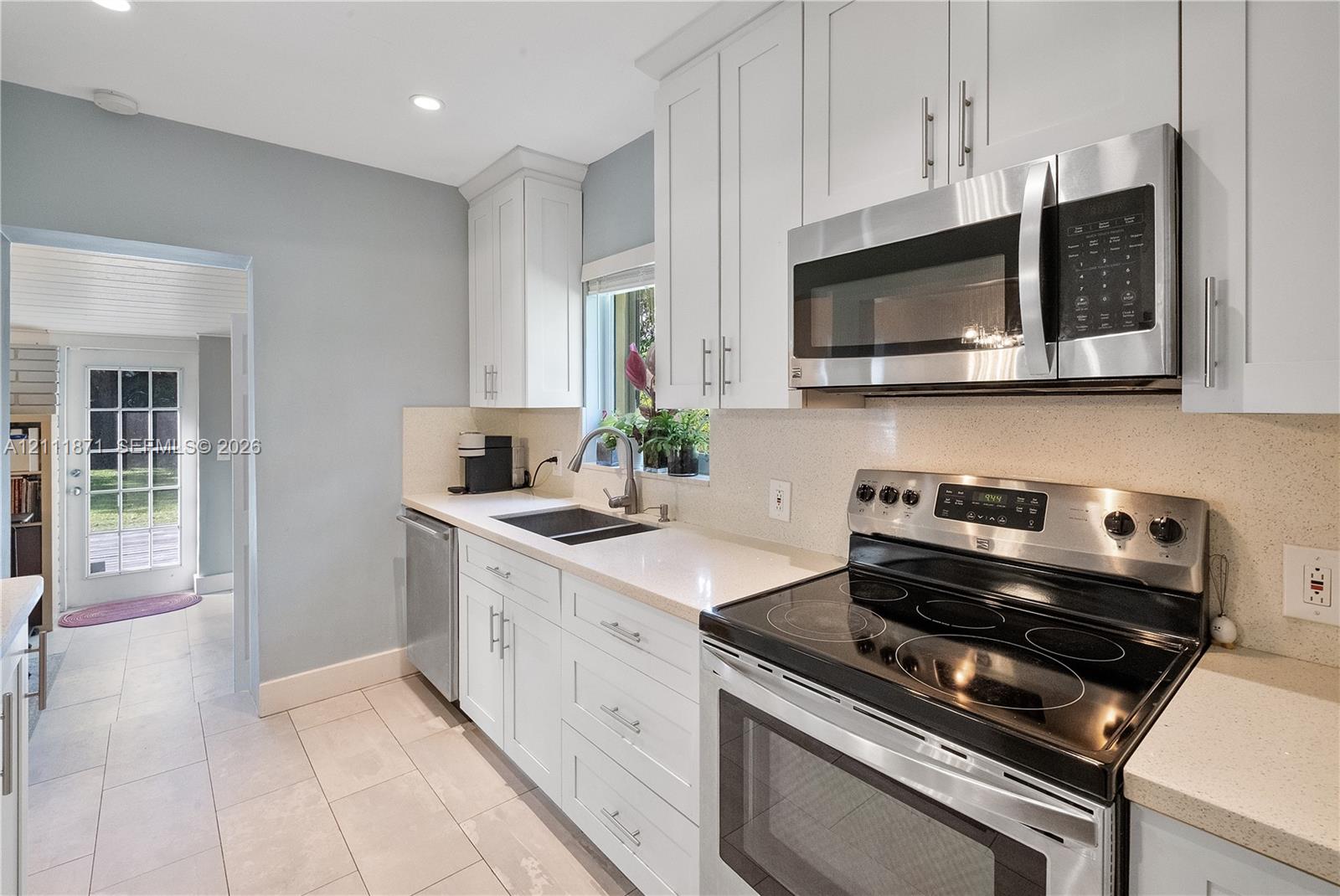 Kitchen featuring white shaker-style cabinetry, light-toned stone countertops, stainless steel appliances, a deep undermount sink, and tiled flooring