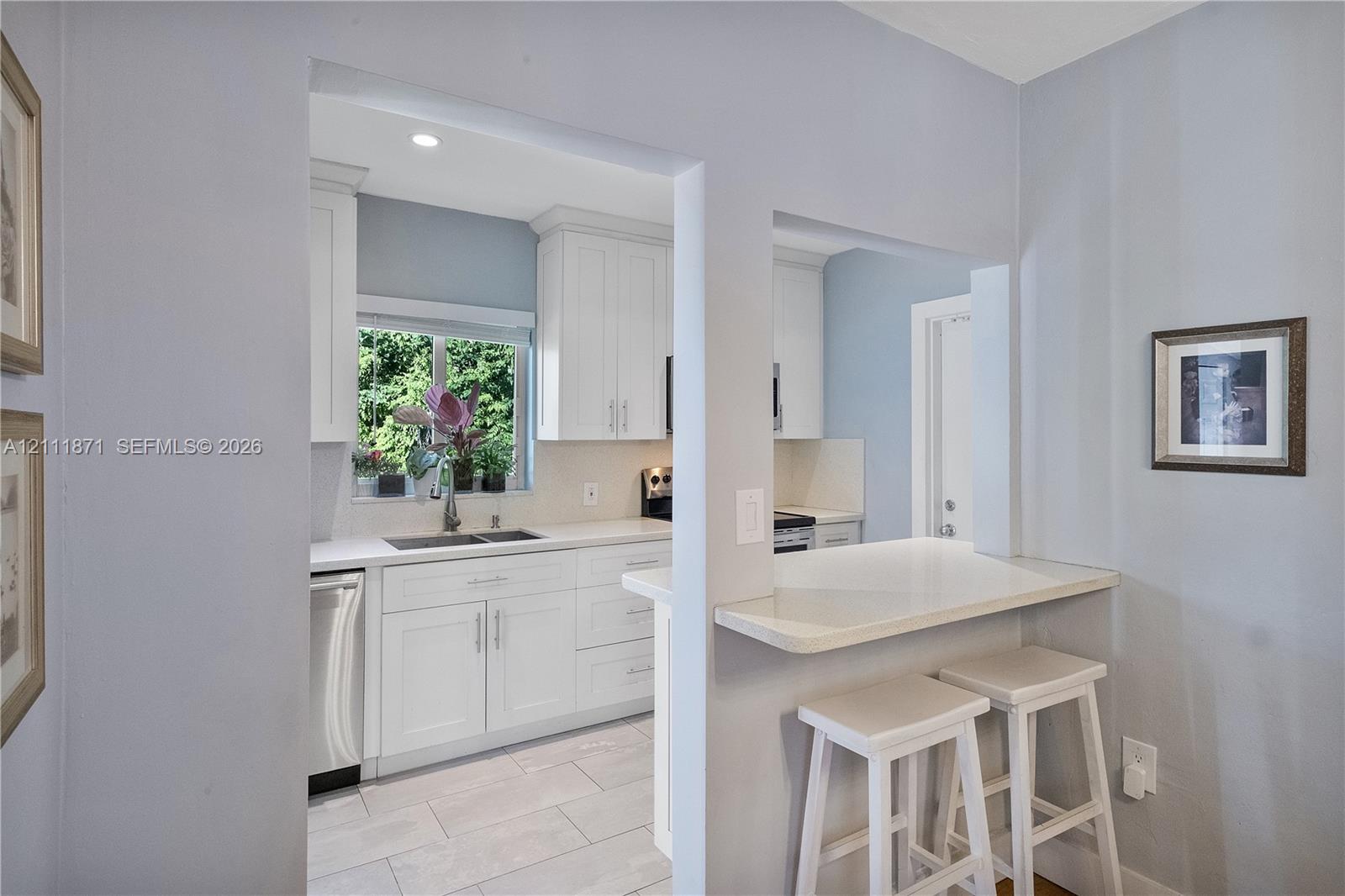 Kitchen featuring white shaker-style cabinetry, solid-surface countertops, dual-basin stainless steel sink, tile flooring, and a built-in pass-through breakfast bar