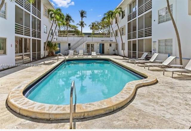 In-ground swimming pool featuring a curved concrete coping, surrounded by a stone paver deck, palm trees, and multi-story structures with screened enclosures
