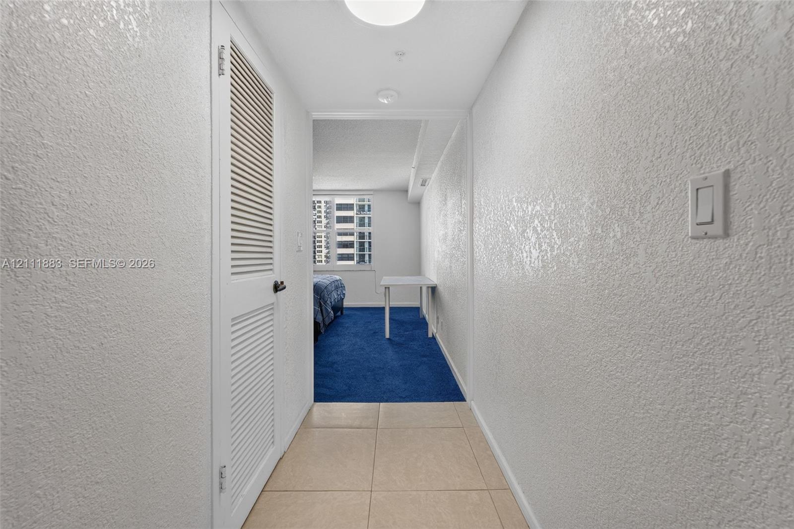 Hallway featuring large-format ceramic tile flooring, textured wall surfaces, and a full-height louvered door closet
