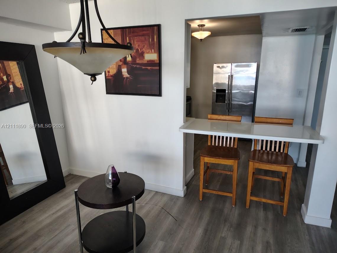 Dining area featuring a modern multi-arm pendant light and wood-finish flooring, complemented by a pass-through breakfast bar with a solid surface countertop and stainless steel refrigerator Dining area featuring a modern multi-arm pendant light and wood-finish flooring, complemented by a pass-through breakfast bar with a solid surface countertop and stainless steel refrigerator