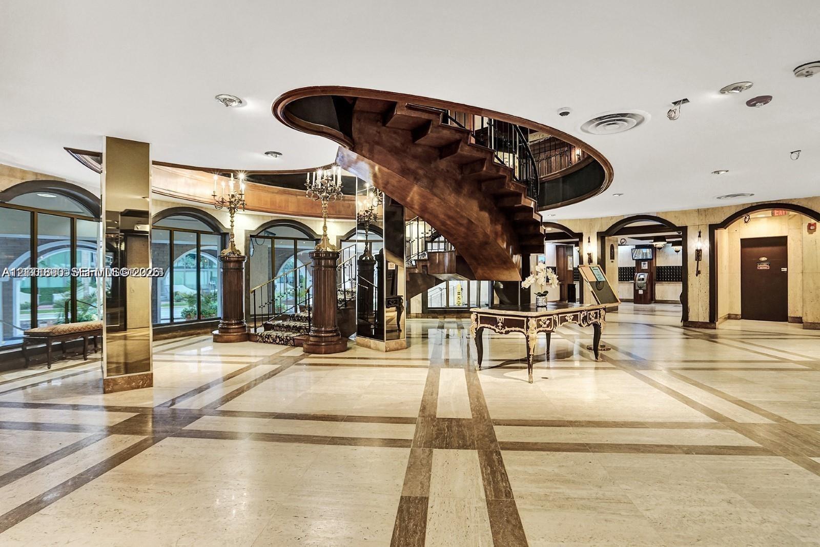 Grand lobby featuring a suspended spiral wooden staircase, polished stone tile flooring with decorative inlay, arched entryways, mirrored support columns, and recessed ceiling lighting