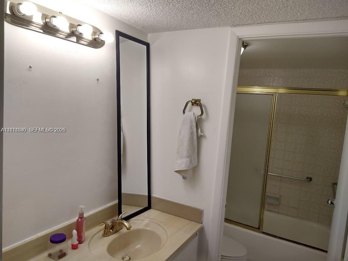 Bathroom featuring a vanity with integrated sink, brass-tone hardware, a vertical rectangular mirror, four-bulb vanity lighting fixture, and a tub-shower combination with a gold-tone sliding frame