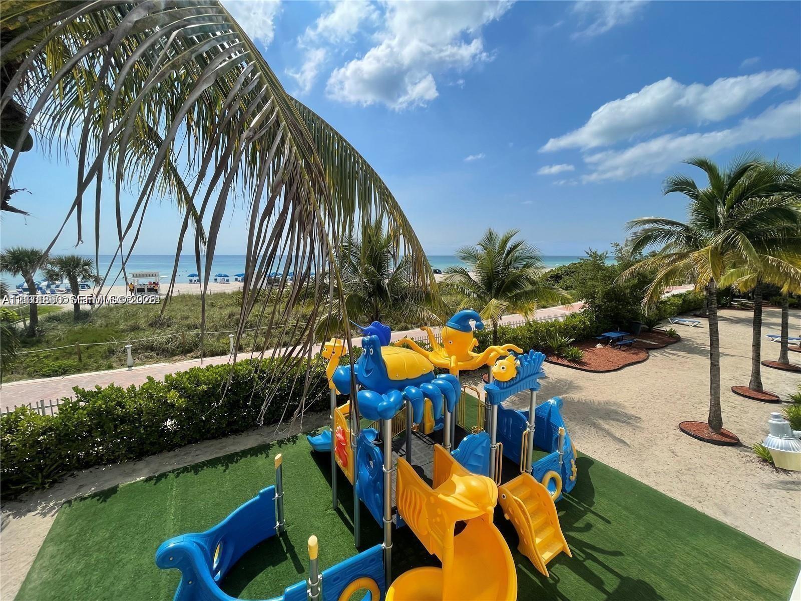 Outdoor playground area featuring artificial turf surfacing, multi-level structures, sand landscaping, mature palm trees, and ocean shoreline proximity