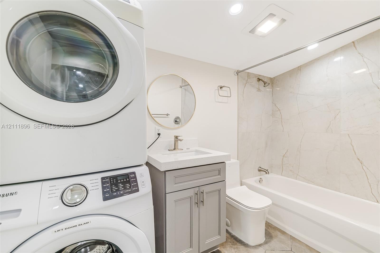Bathroom featuring a vanity with a white countertop and grey cabinetry, a round wall-mounted mirror, a tub-shower combination with full-height tile surround, and a stacked laundry unit