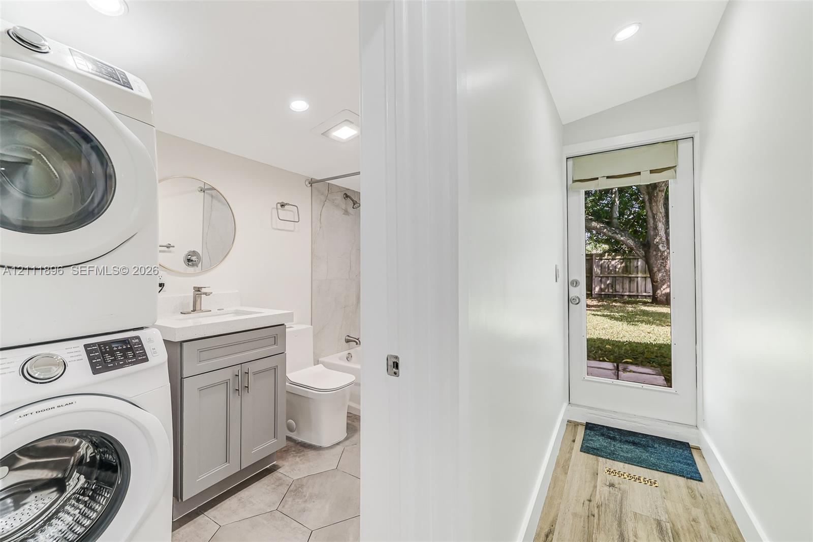 Combined utility and bathroom featuring a stacked washer and dryer, gray vanity with a rectangular basin, tile flooring, and exterior door with glass insert