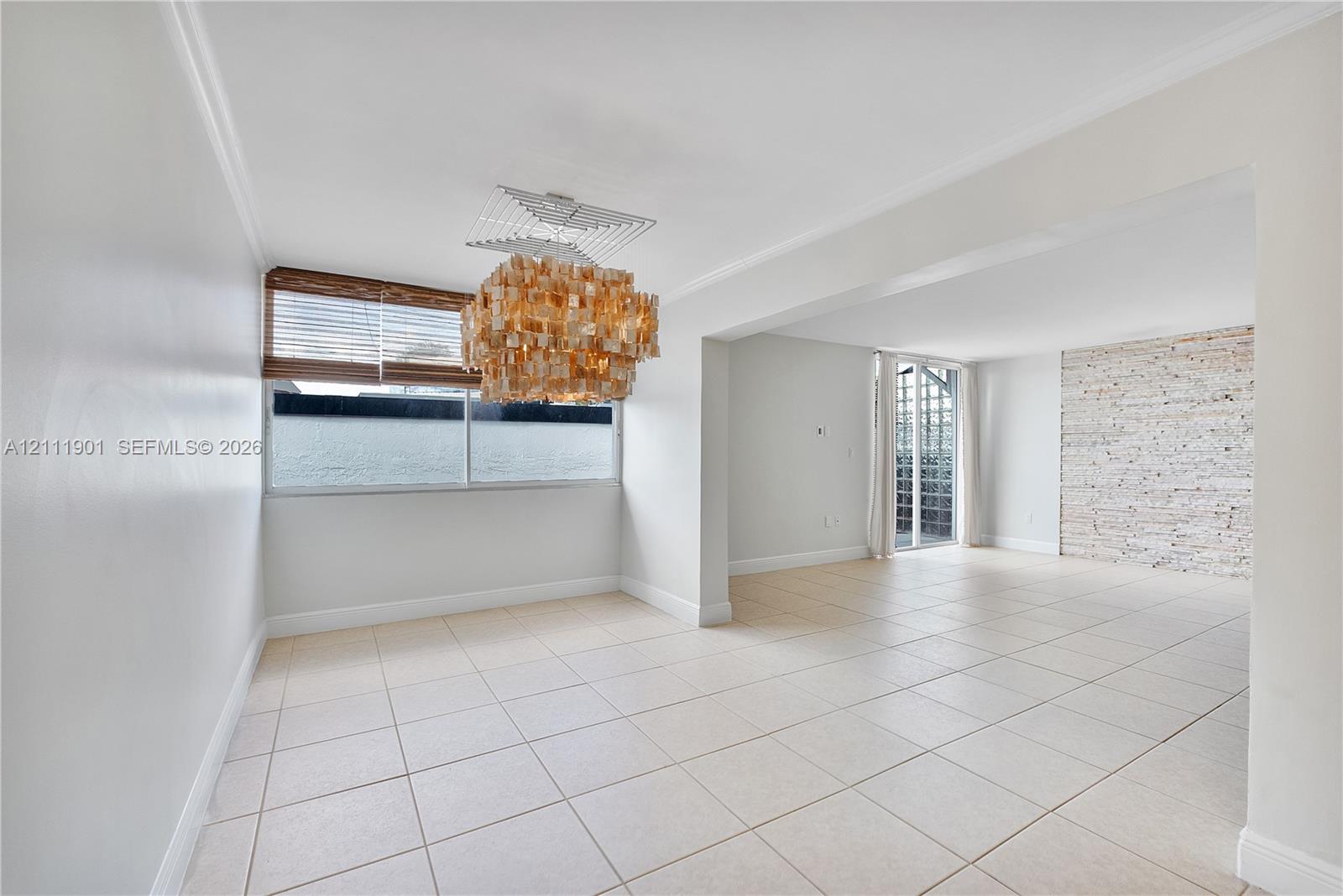 Open interior featuring large-format tile flooring, textured stone accent wall, multi-tiered crystal statement light fixture, and horizontal blind window coverings