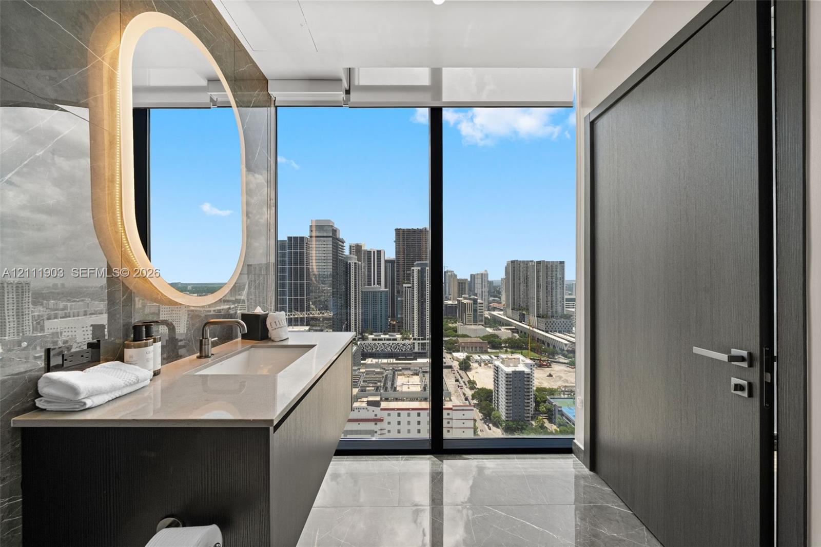 Floating vanity featuring a stone countertop, integrated sink, and wall-mounted faucet, complemented by an oval backlit mirror, full-height window, and polished stone flooring
