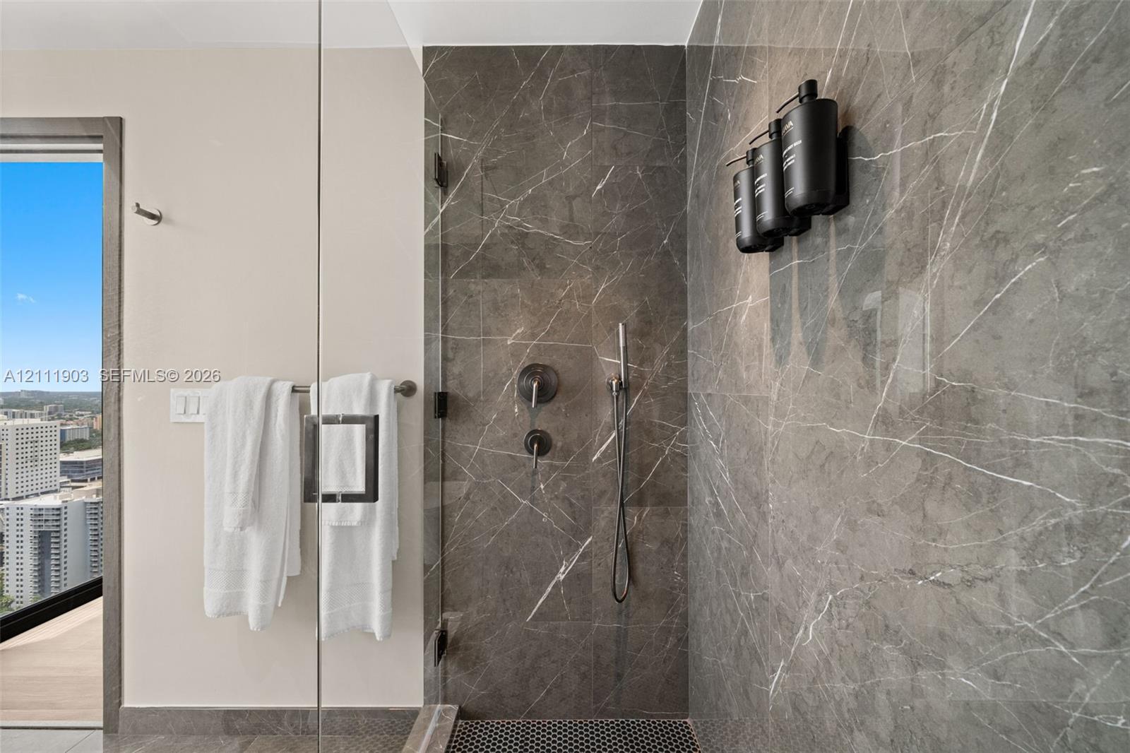 Walk-in shower featuring floor-to-ceiling stone-finish tile, frameless glass enclosure, dark-finish wall-mounted shower hardware, and mosaic tile flooring