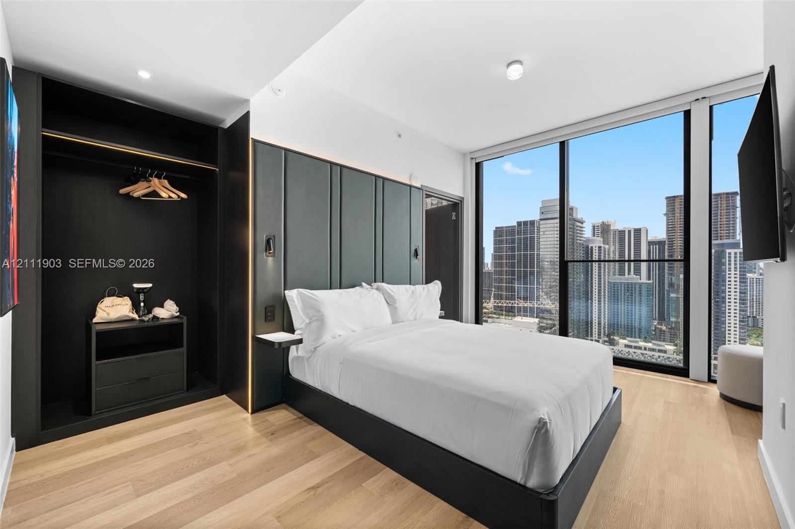 Floor-to-ceiling windows with urban skyline exposure, wood-finish flooring, built-in open-concept wardrobe with integrated storage, and decorative wall paneling with mounted accent lighting