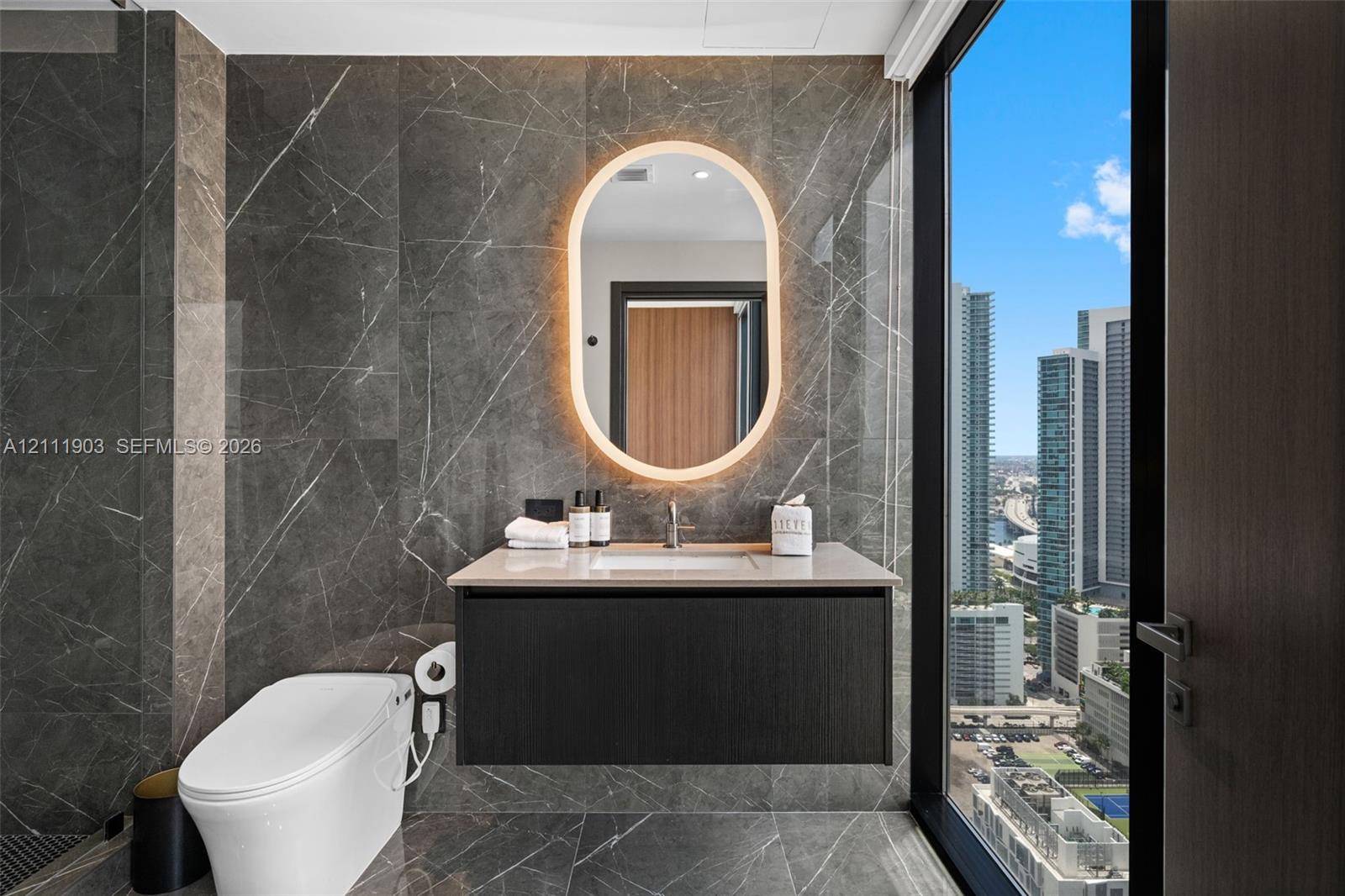 Full-height stone wall tiling, modern wall-mounted vanity with a stone countertop, oval LED-backlit mirror, floor-to-ceiling windows, and wall-mounted toilet