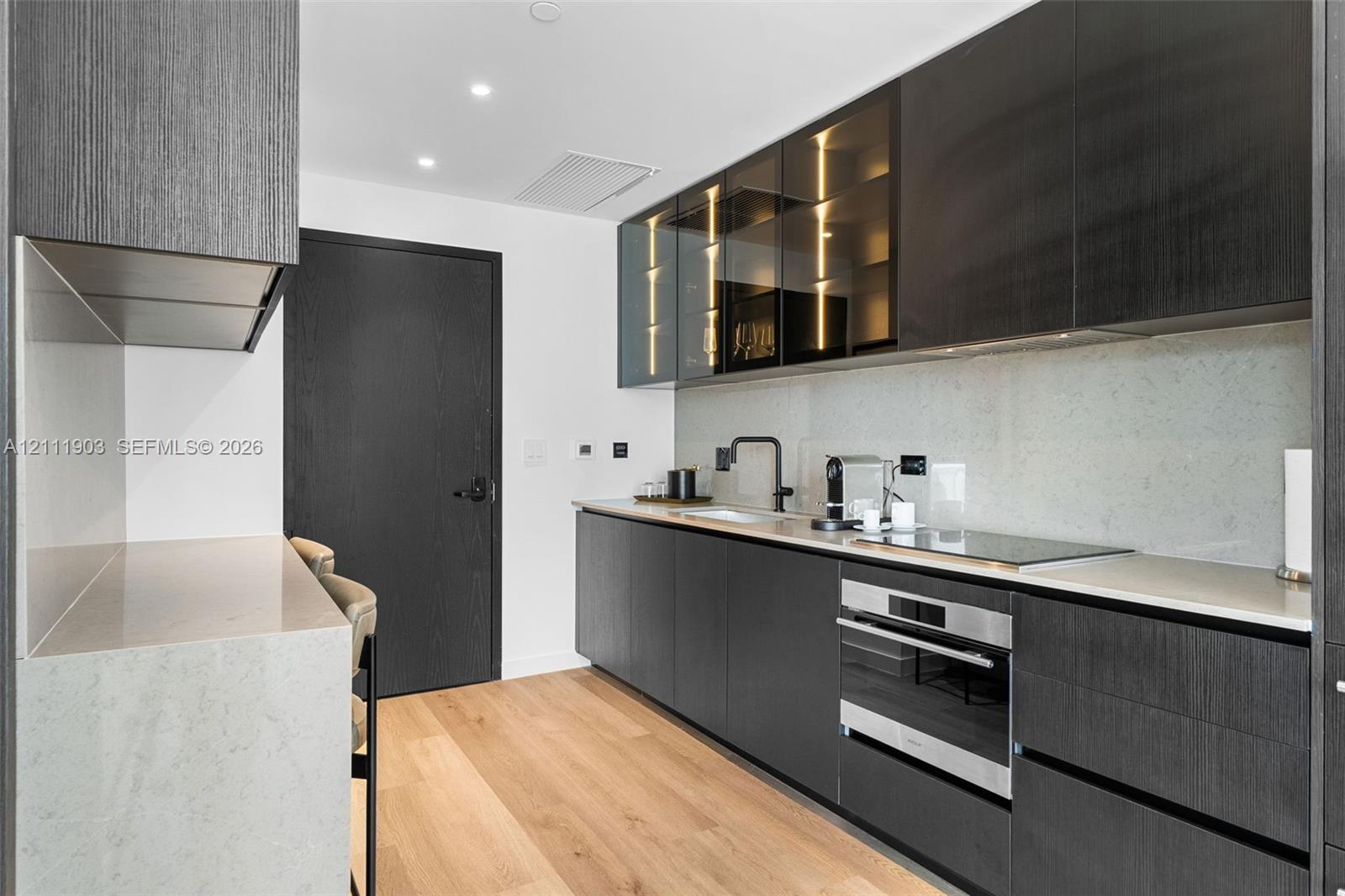 Modern kitchen featuring flat-panel dark cabinetry, stone-finish countertops, glass-front upper cabinets, a built-in stainless steel oven, and wood-finish flooring