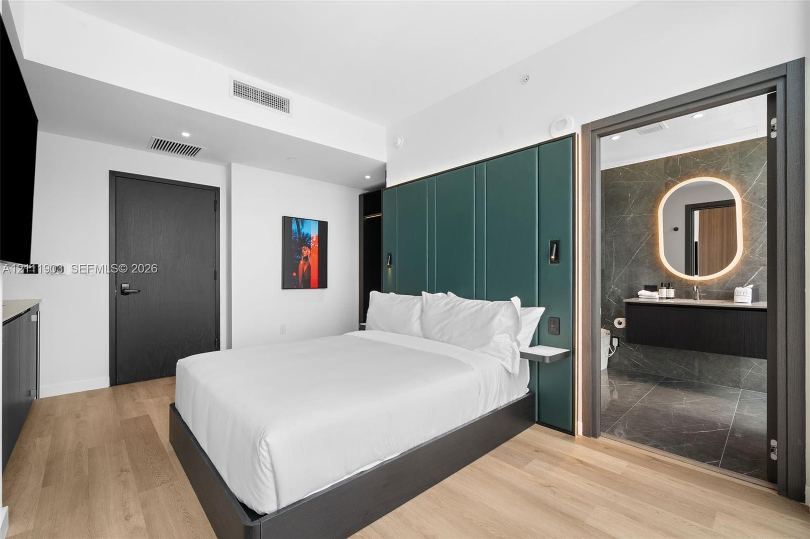 Custom floor-to-ceiling green panel headboard, wood-finish flooring, dark-toned interior doors, floating bathroom vanity, and rounded illuminated mirror