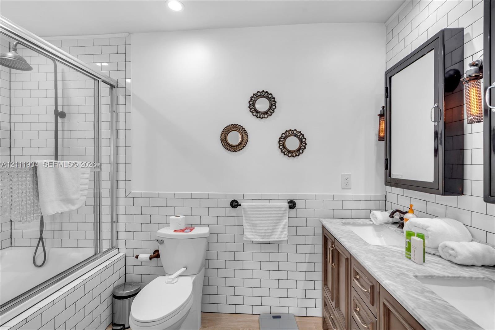 Bathroom featuring white subway tile wall treatments, a dual-sink vanity with light-toned stone-finish countertop, industrial-style wall-mounted medicine cabinets, and a glass-enclosed shower with a rain-style showerhead