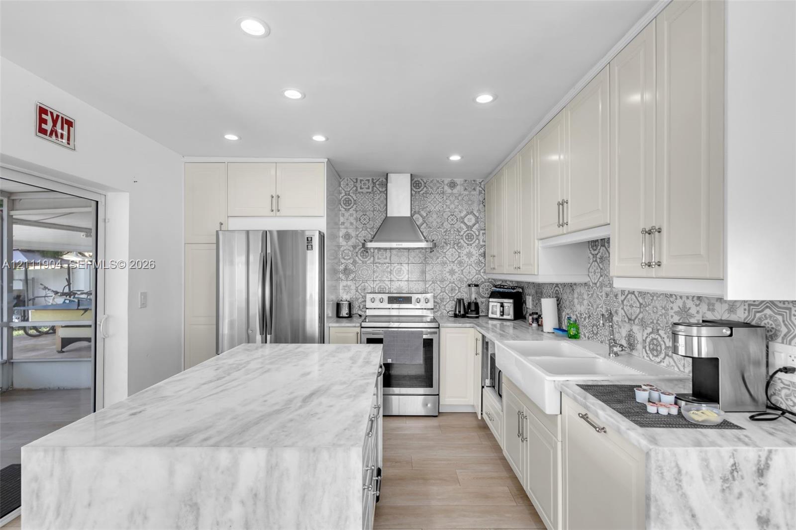 Kitchen featuring stone-topped central island, patterned tile backsplash, apron-front sink, stainless steel appliances, and recessed lighting