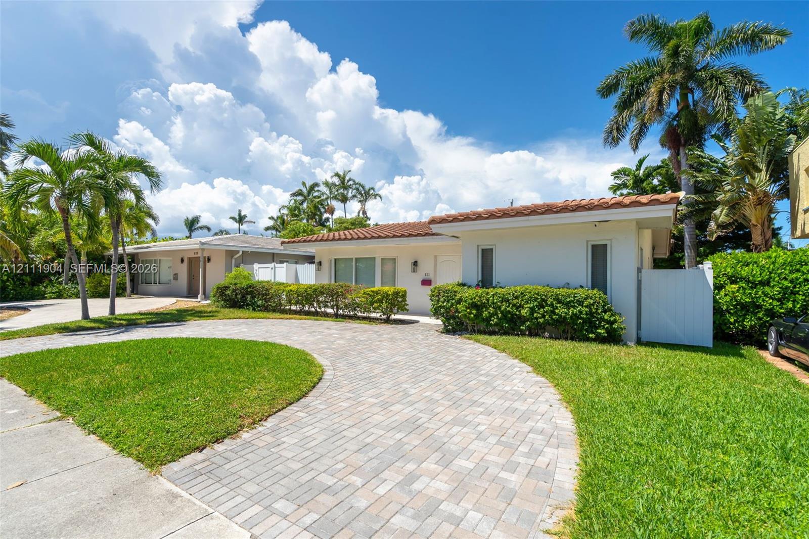 Single-story exterior featuring a clay tile roof, white stucco finish, circular paver driveway, and manicured lawn with mature palm trees