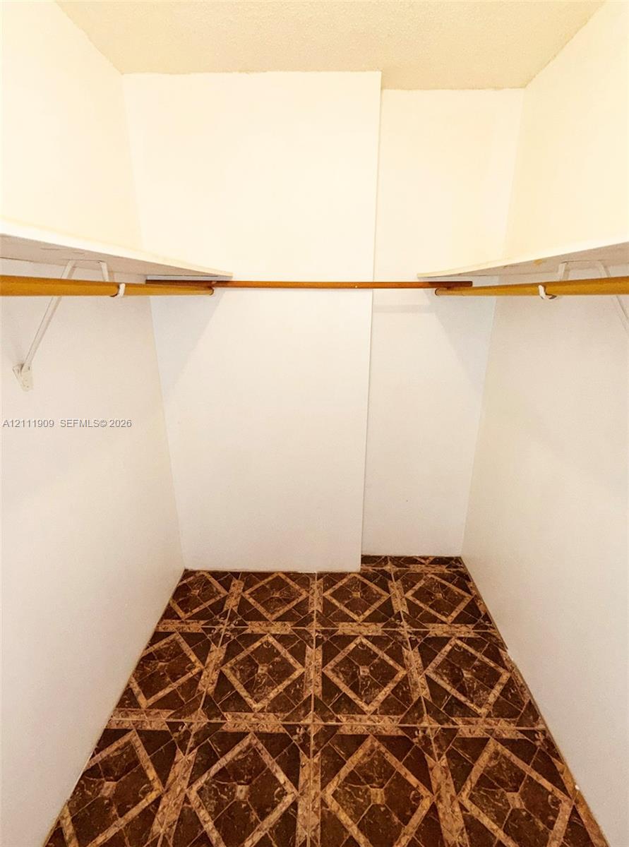 Storage space featuring white walls, upper shelving, and installed hanging rods over geometric-patterned tile flooring