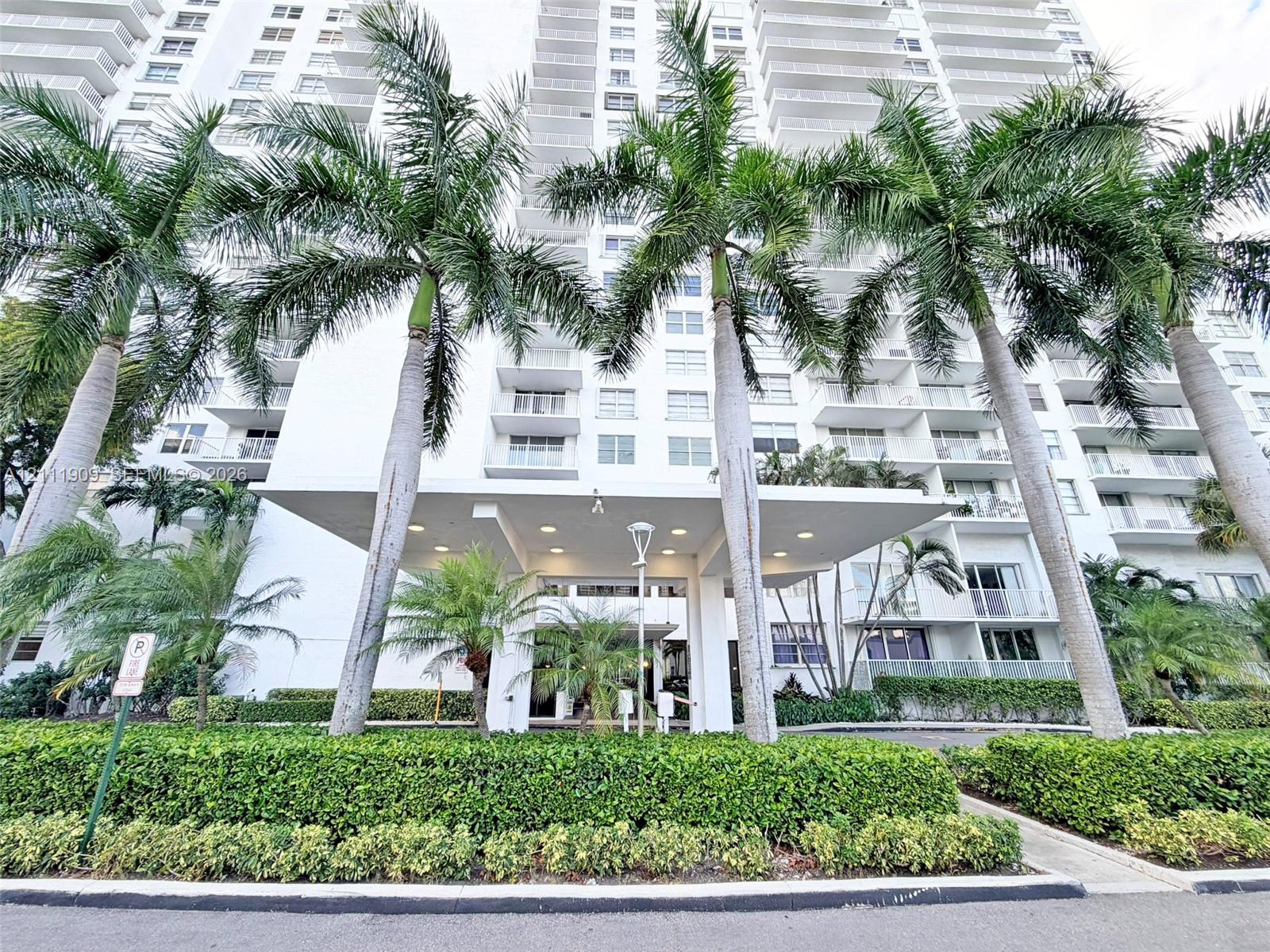 High-rise building exterior featuring a covered porte-cochere entry, wrap-around private balconies, recessed lighting, and manicured landscaping with mature palm trees
