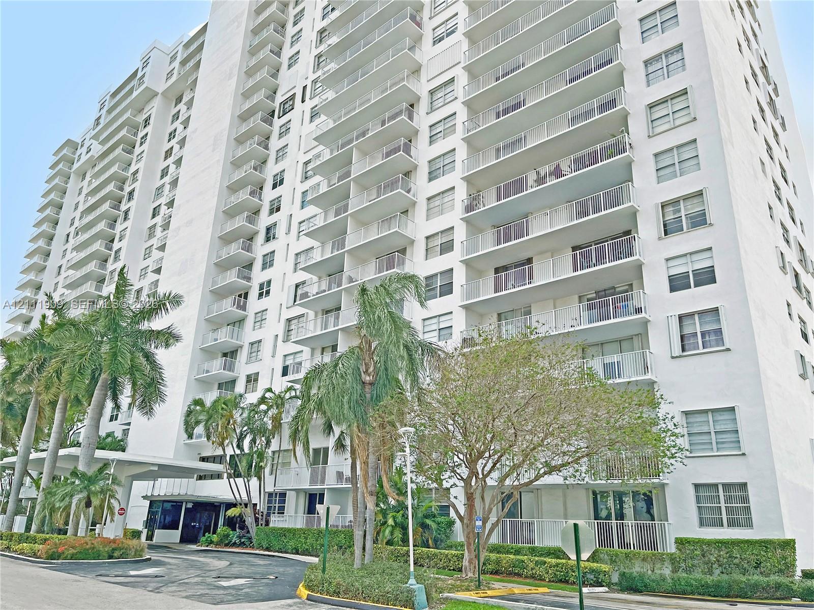 High-rise multi-story building featuring tiered private balconies, an expansive concrete motor court, a covered entryway portico, and mature tropical landscaping
