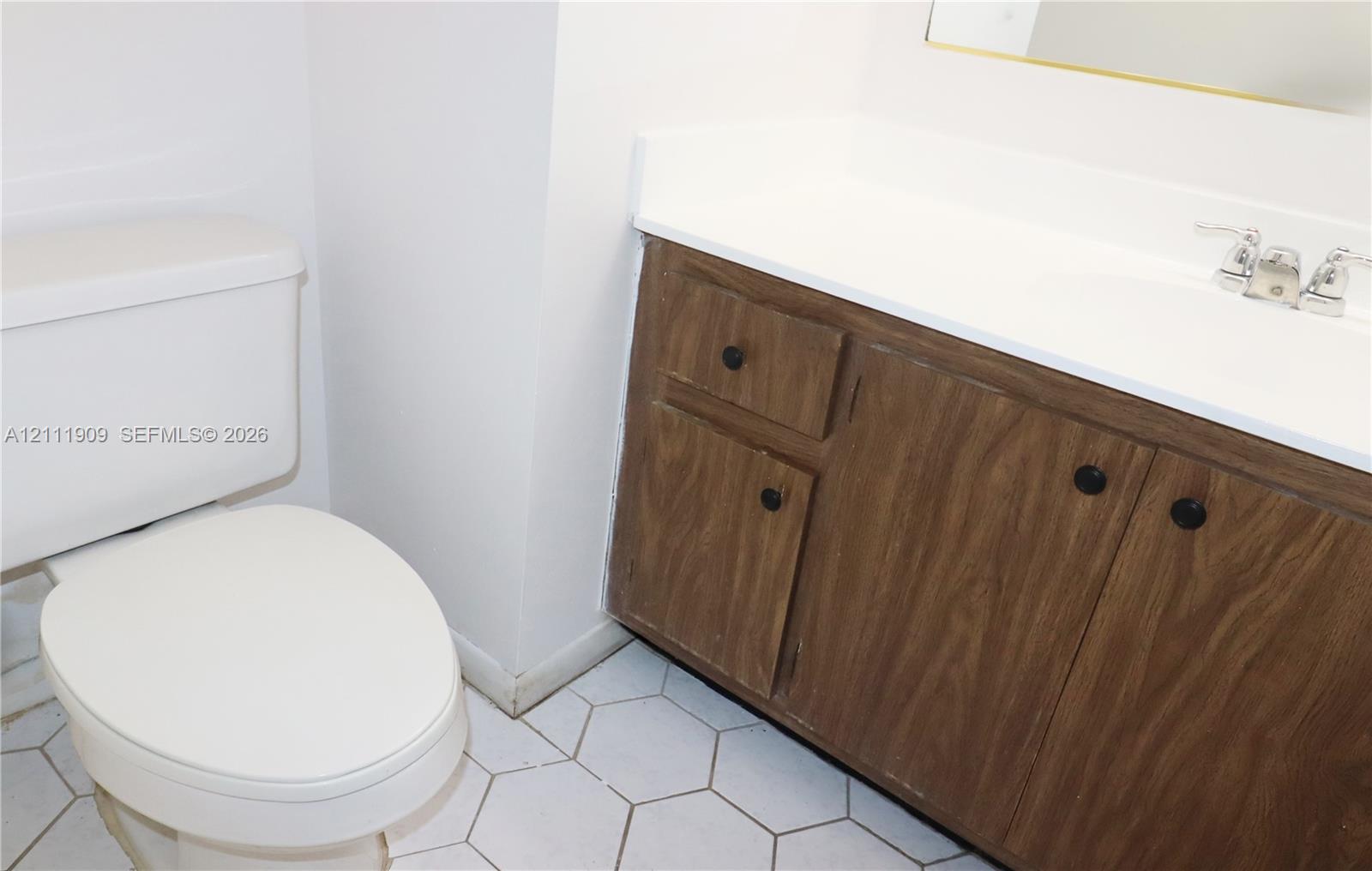 Bathroom featuring a wood-finish vanity with a white countertop, integrated sink, chrome dual-handle faucet, and white hexagonal floor tiling