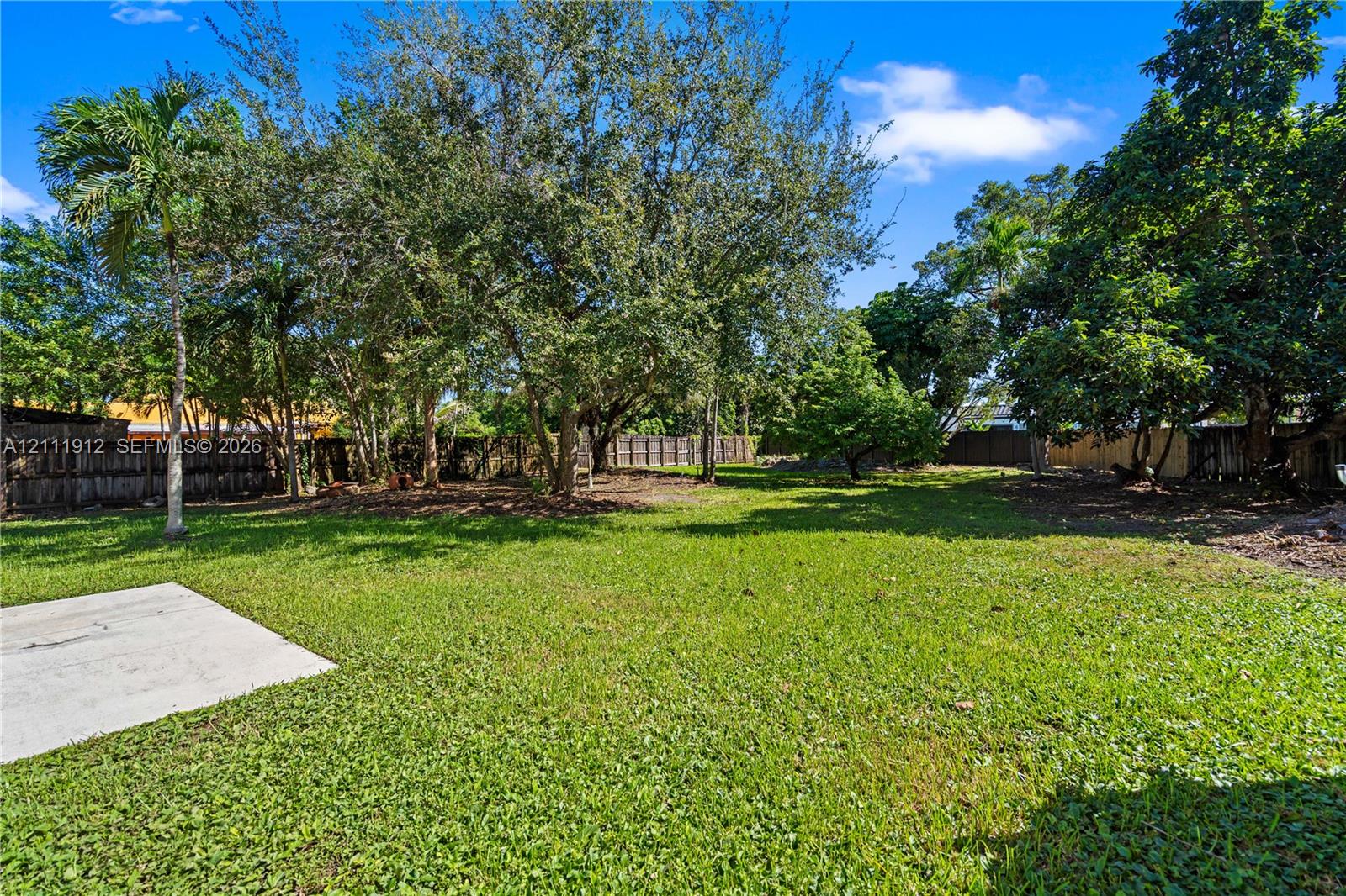 Expansive backyard featuring a concrete patio, mature trees, and wood-panel perimeter fencing