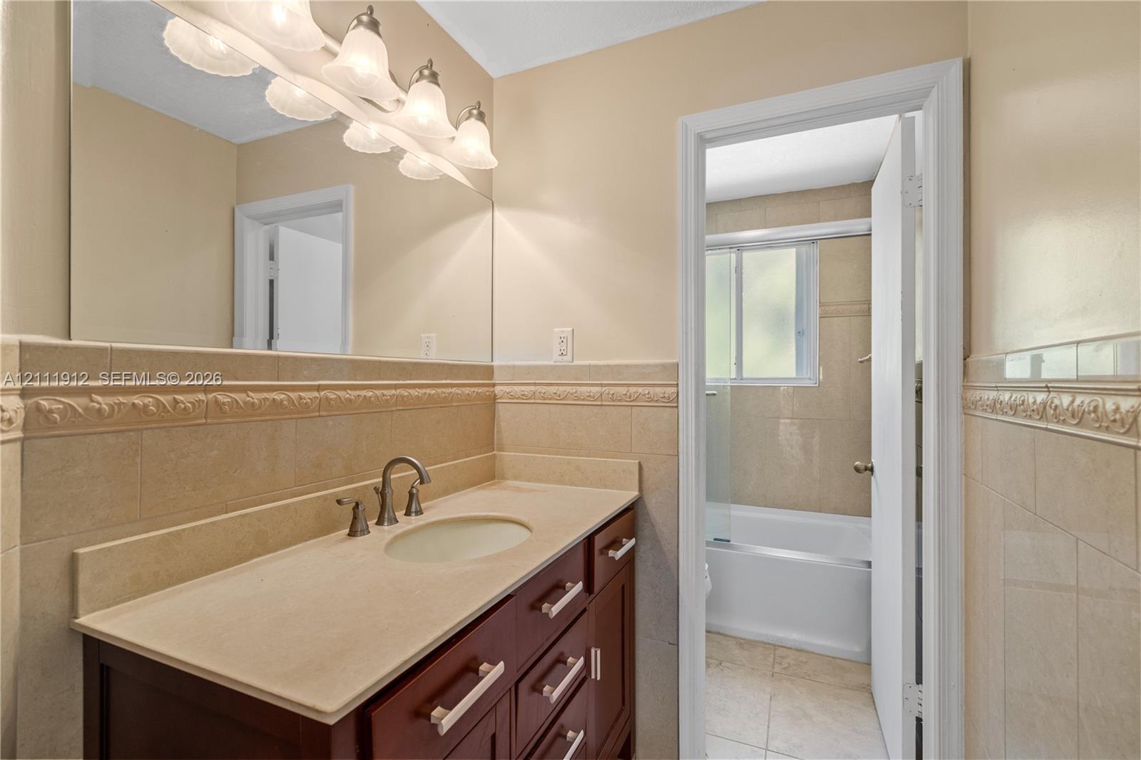 Bathroom featuring a dark wood-finish vanity, stone countertop with an integrated sink, full-width wall mirror, and neutral wall tiling with decorative relief trim