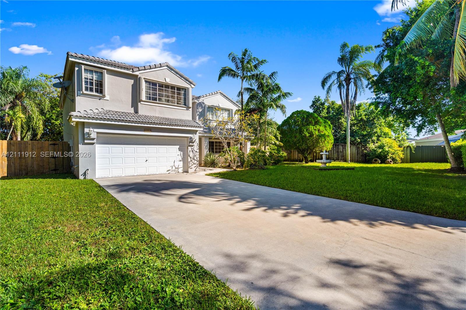 Two-story exterior featuring a light-toned stucco faade, tile roof, multi-car garage, concrete driveway, and expansive lawn with mature palm trees