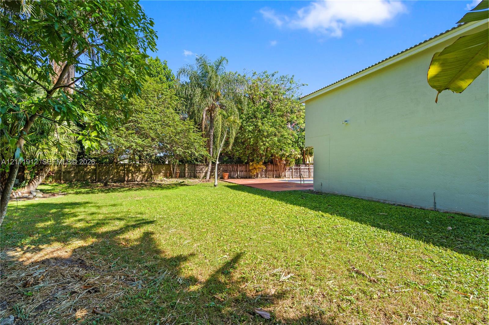 Spacious backyard featuring a large grass lawn, perimeter wood fencing, mature trees, and a paved patio area
