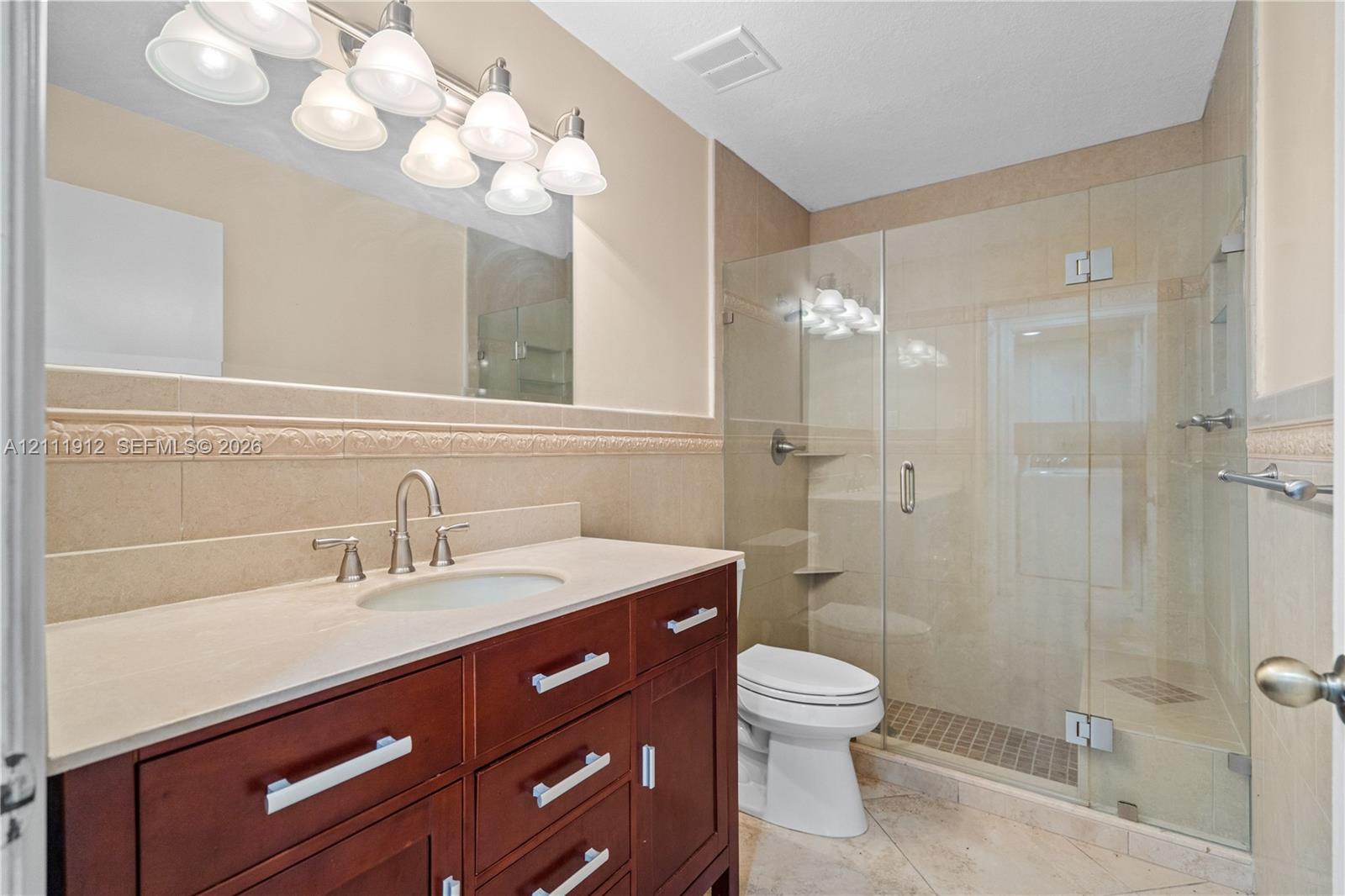 Bathroom featuring a dark wood-finish vanity, light-toned countertop with an integrated sink, frameless glass-enclosed shower, decorative tile wall border, and multi-bulb vanity lighting