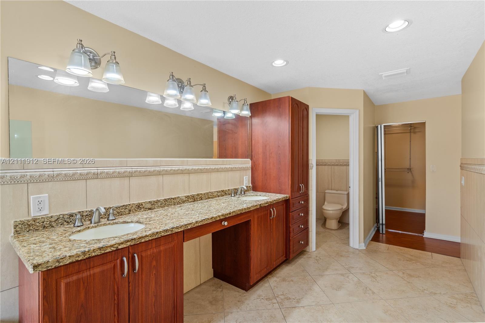 Bathroom featuring a dual-sink vanity with stone countertop, wood-finish cabinetry, full-width wall mirror, and tile flooring