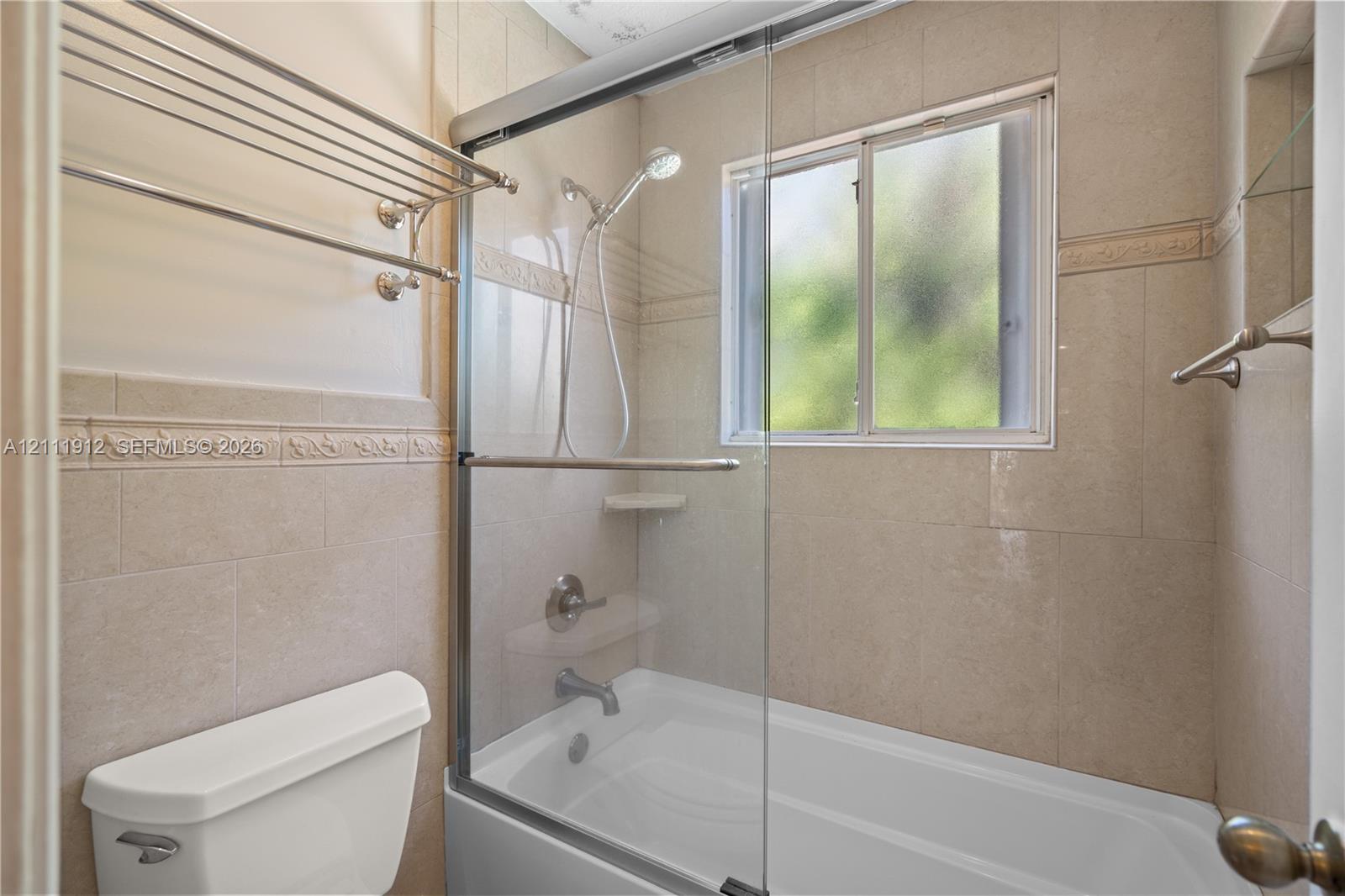 Tub and shower combination featuring floor-to-ceiling tile surround with decorative inlay, glass sliding enclosure, handheld shower fixture, and wall-mounted metal shelving