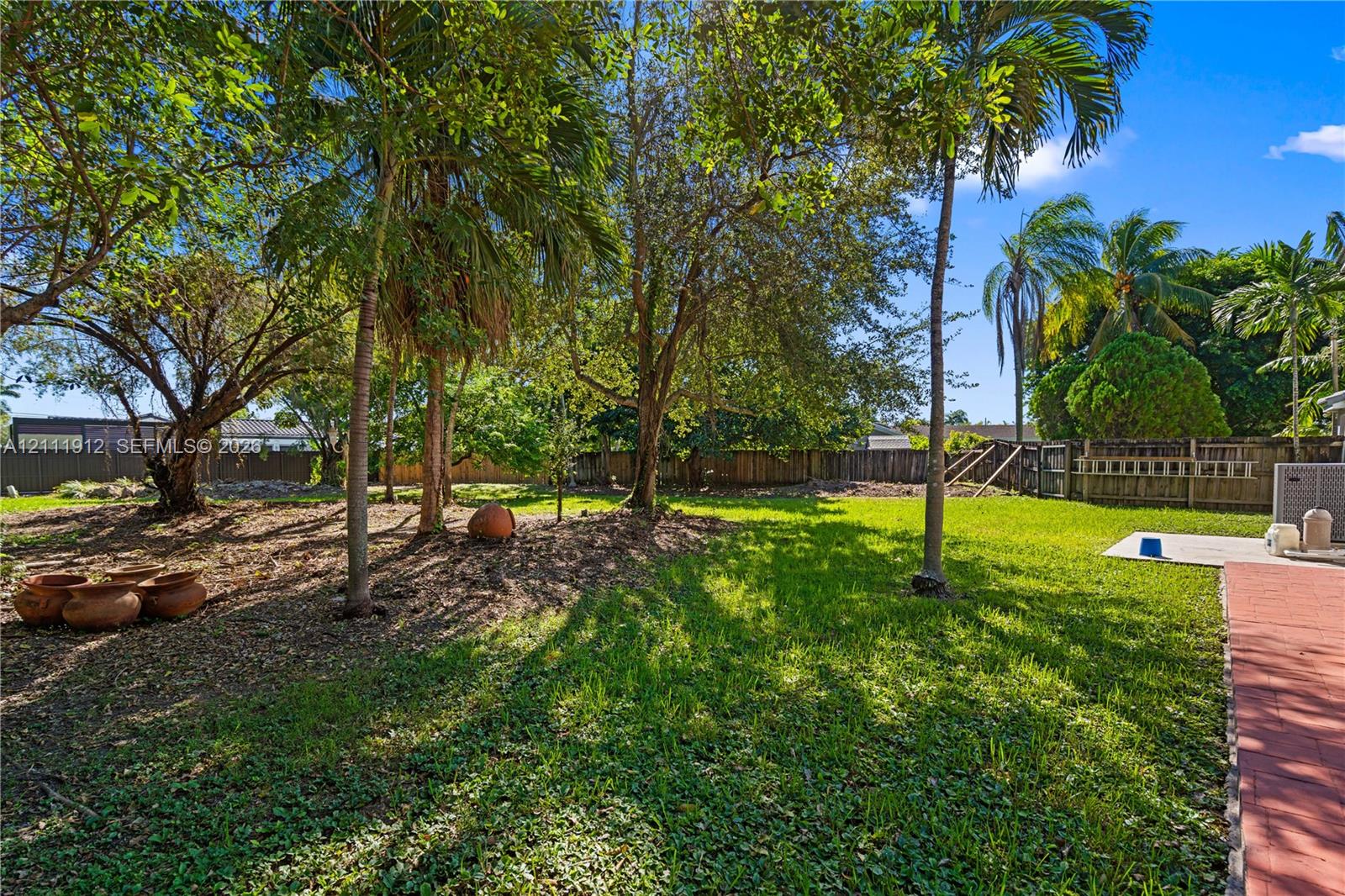 Expansive backyard featuring mature palm trees, established landscaping, perimeter wood fencing, and brick-paver patio surfaces