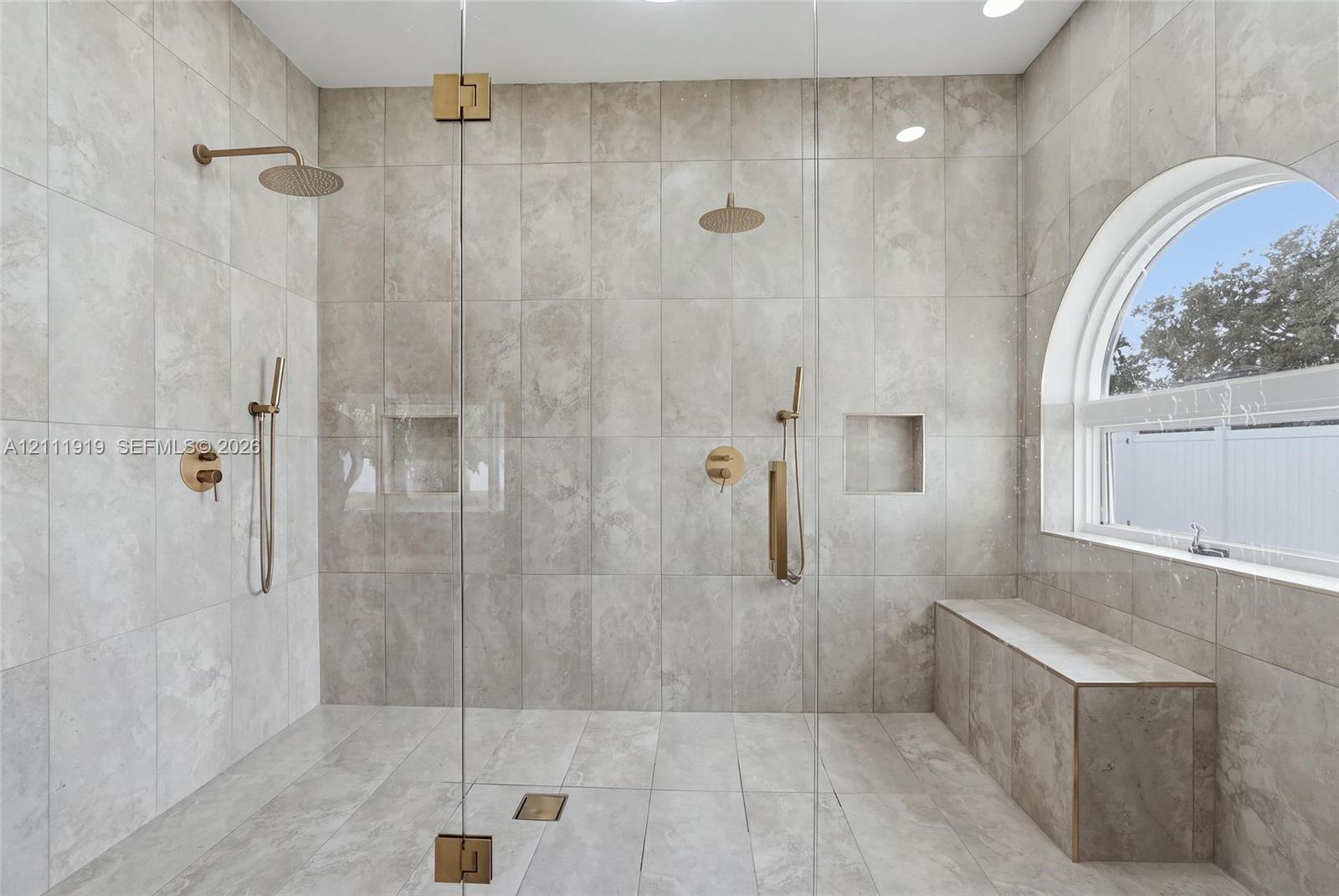 Walk-in shower featuring floor-to-ceiling large-format neutral tile, dual rainfall showerheads with handheld sprayers, built-in recessed niches, a tiled bench, and a signature arched window