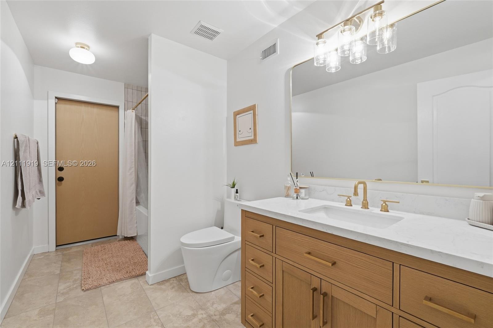 Bathroom featuring a wood-finish vanity with a stone-surface countertop, rectangular undermount sink, brass-tone fixtures, oversized wall-to-wall mirror, and neutral tile flooring