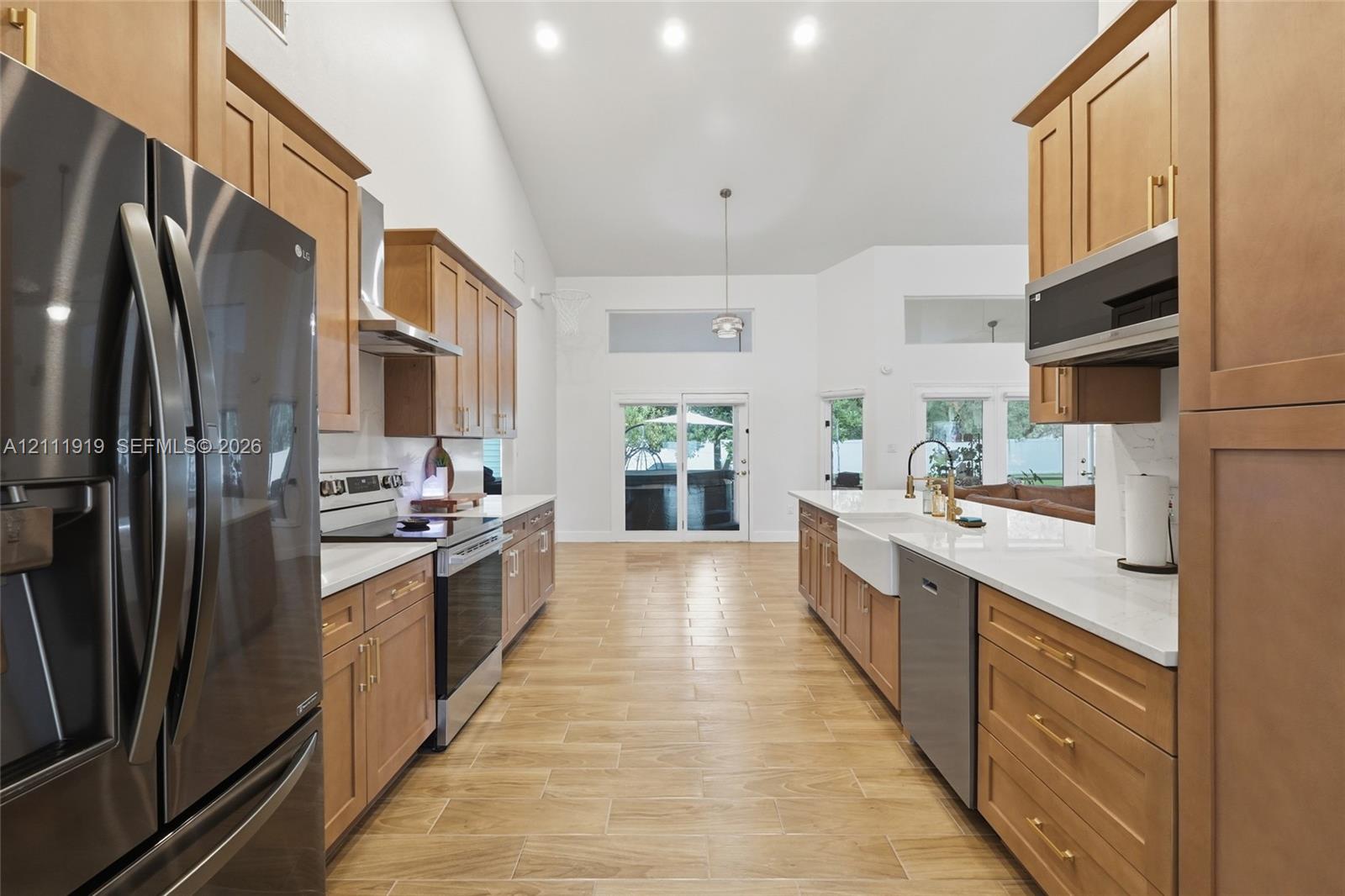 Kitchen featuring light wood cabinetry with gold-tone hardware, white countertops, a farmhouse-style sink, stainless steel appliances, and wood-finish tile flooring under a vaulted ceiling Kitchen featuring light wood cabinetry with gold-tone hardware, white countertops, a farmhouse-style sink, stainless steel appliances, and wood-finish tile flooring under a vaulted ceiling