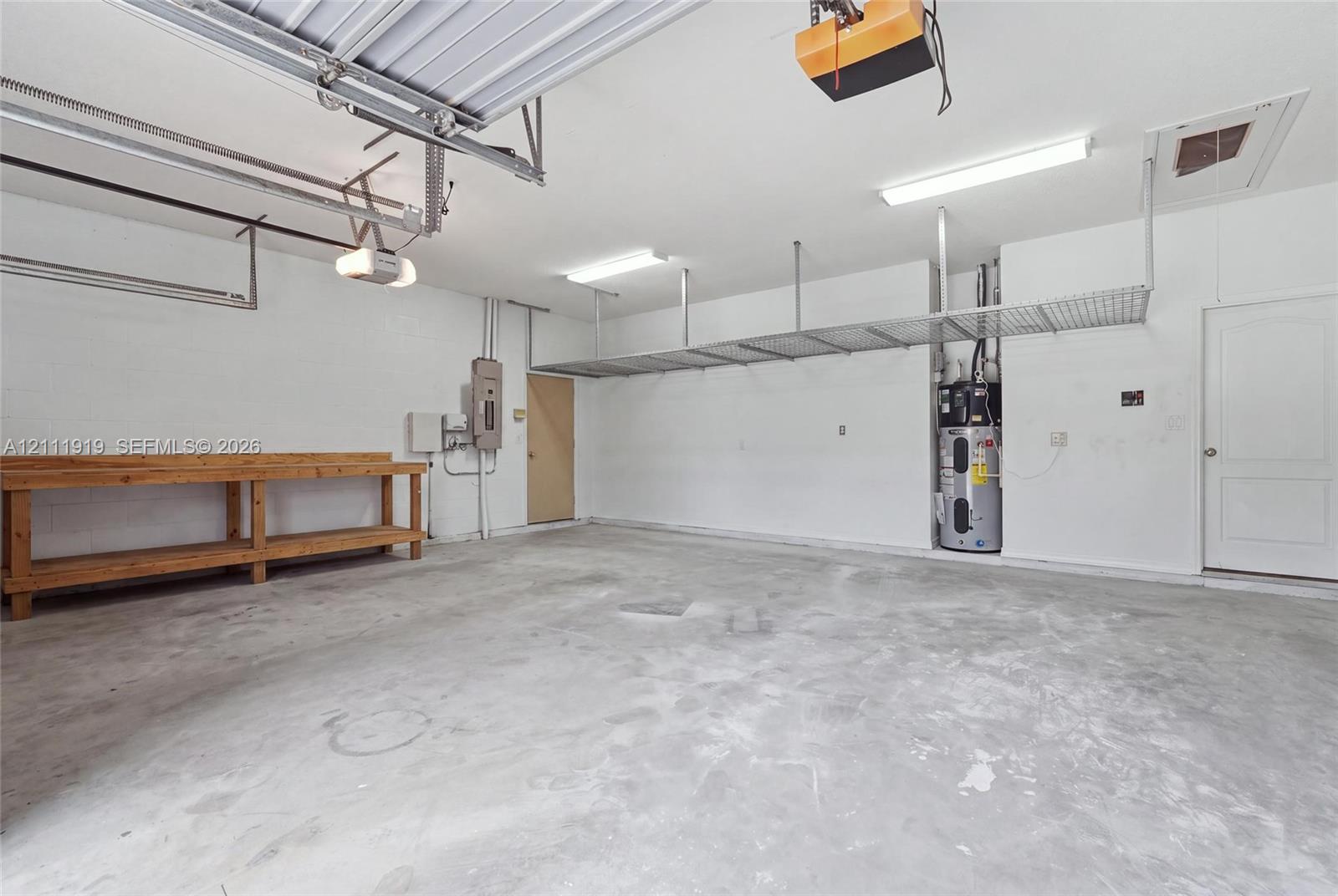 Spacious garage featuring concrete flooring, built-in wooden workbench, overhead metal storage shelving, and dedicated water heater enclosure