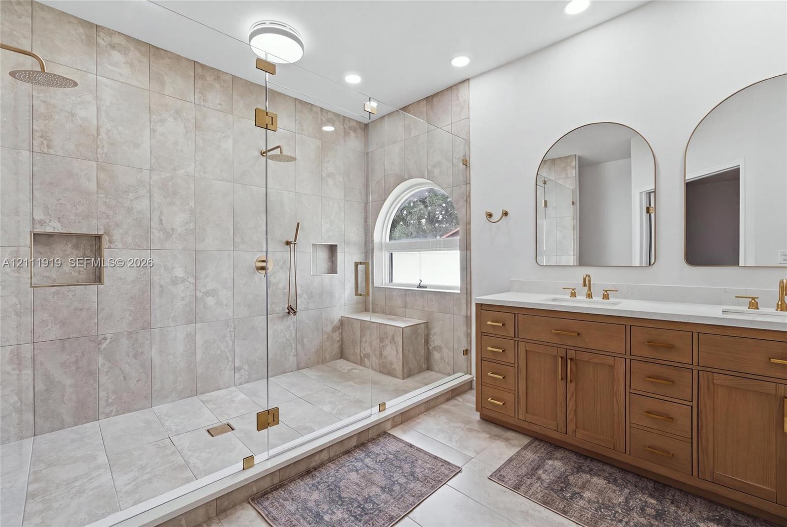 Walk-in shower featuring floor-to-ceiling stone-finish tile, an integrated bench, and a glass enclosure, paired with a dual-sink vanity with brass-finish hardware and arched mirrors