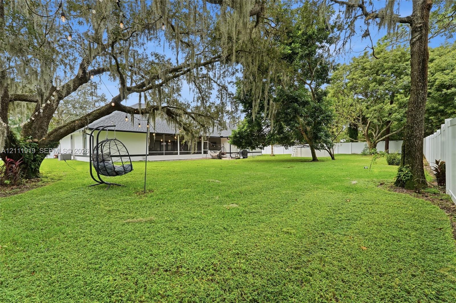 Spacious backyard featuring a manicured lawn, mature trees, white privacy fencing, and a screened-in lanai