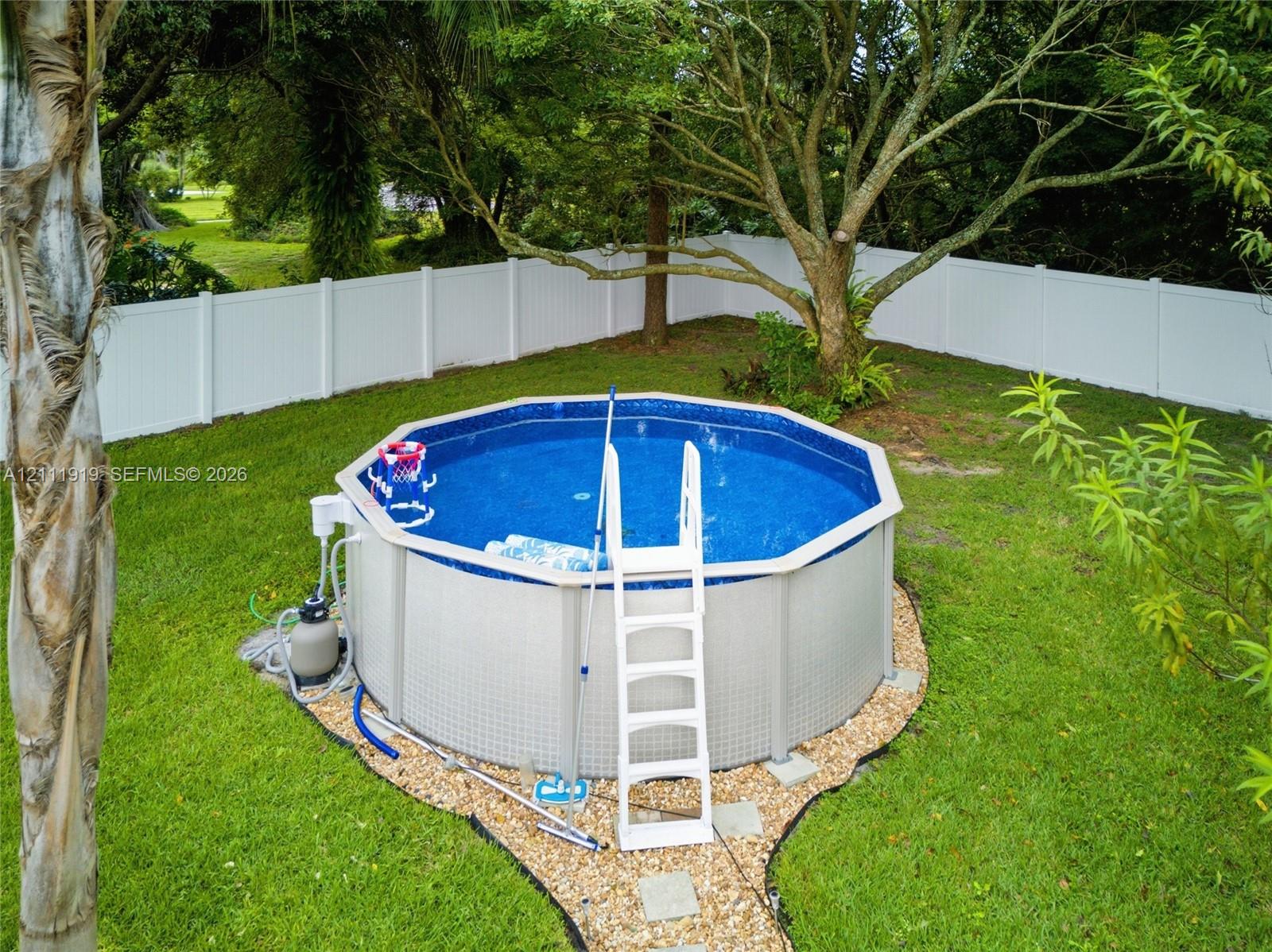 Above-ground swimming pool with surrounding gravel border, enclosed by a white vinyl privacy fence and set within a grass-covered backyard featuring mature trees Above-ground swimming pool with surrounding gravel border, enclosed by a white vinyl privacy fence and set within a grass-covered backyard featuring mature trees