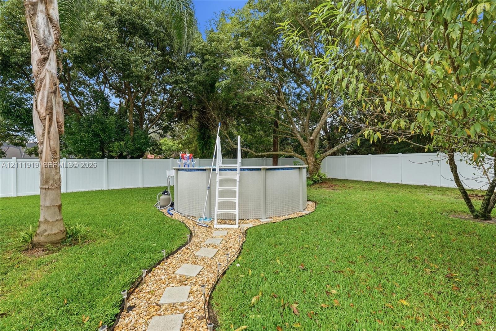 Backyard featuring an above-ground swimming pool, white vinyl fencing, a gravel path with stone pavers, and mature trees