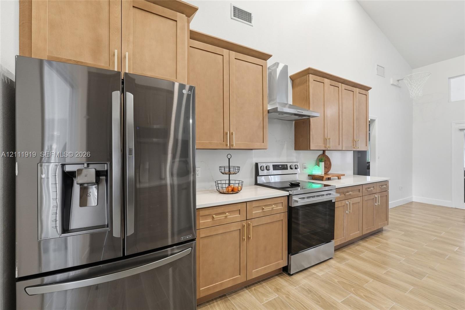 Kitchen featuring wood-finish cabinetry, light-toned countertops, stainless steel appliances, a wall-mounted chimney-style range hood, and wood-finish flooring Kitchen featuring wood-finish cabinetry, light-toned countertops, stainless steel appliances, a wall-mounted chimney-style range hood, and wood-finish flooring