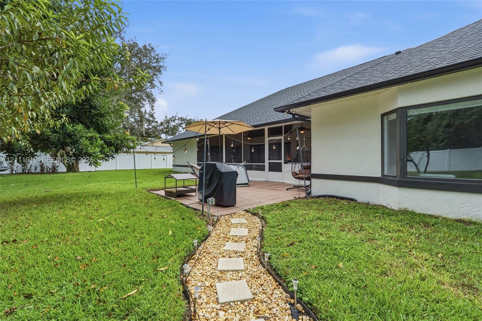 Rear exterior featuring a screened-in lanai, concrete paver patio, gravel walkway with square stone steppers, and a white vinyl perimeter fence Rear exterior featuring a screened-in lanai, concrete paver patio, gravel walkway with square stone steppers, and a white vinyl perimeter fence