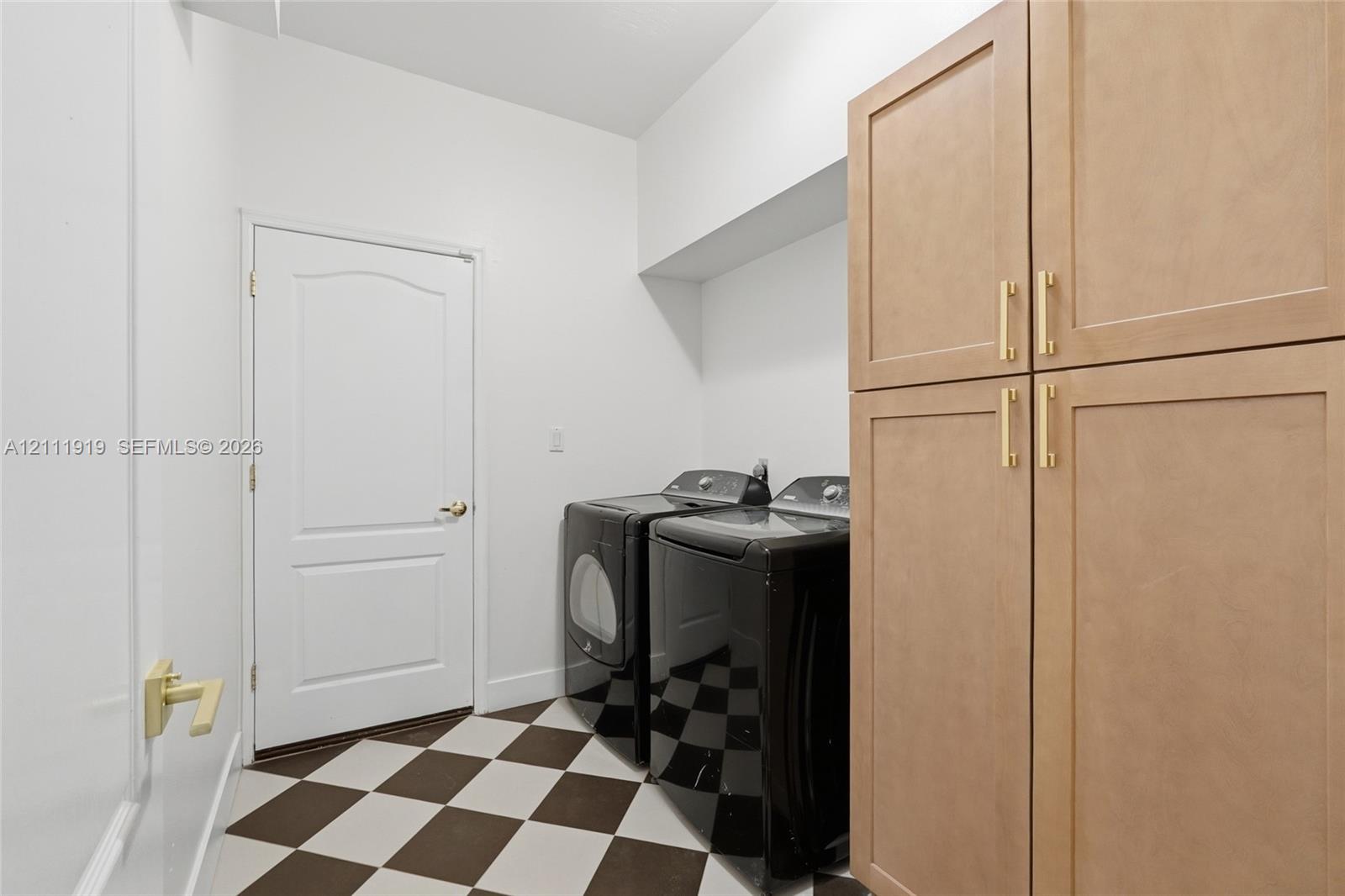 Laundry area featuring built-in light wood cabinetry with gold-toned hardware, a checkerboard tile floor, and a solid-core door with a gold-toned lever handle