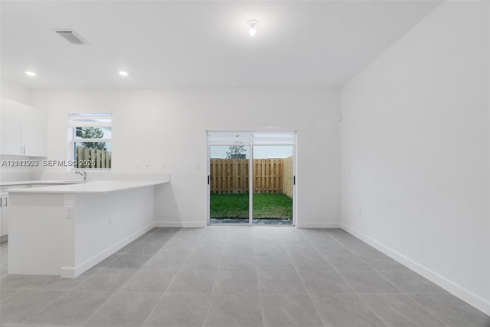 Open-concept interior featuring large-format ceramic tile flooring, white cabinetry, quartz countertop kitchen peninsula, and sliding glass doors with direct access to the fenced backyard