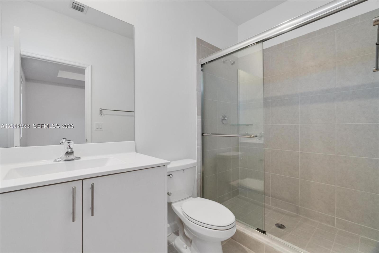 Bathroom featuring a white vanity cabinet with a solid-surface countertop, an integrated sink with a chrome faucet, a large frameless mirror, and a walk-in shower with sliding glass doors and tiled wall surround
