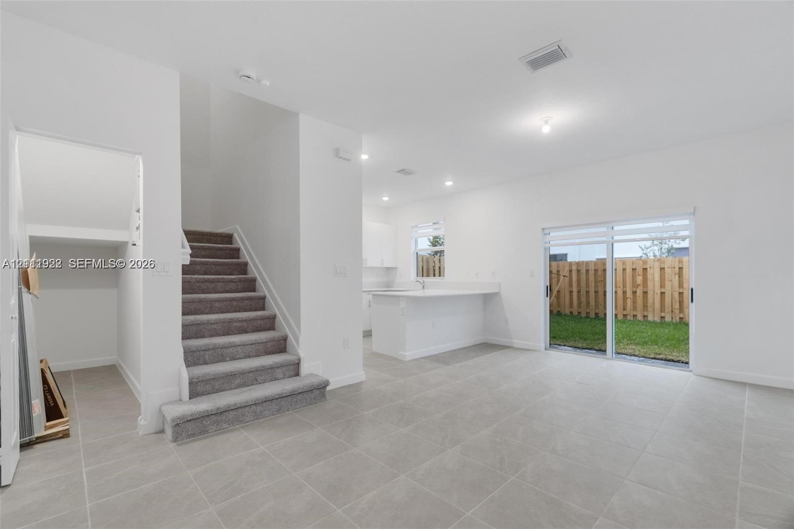 Open-concept interior featuring large-format neutral tile flooring, carpeted staircase, kitchen with white cabinetry and breakfast bar, and sliding glass doors with access to a fenced rear yard