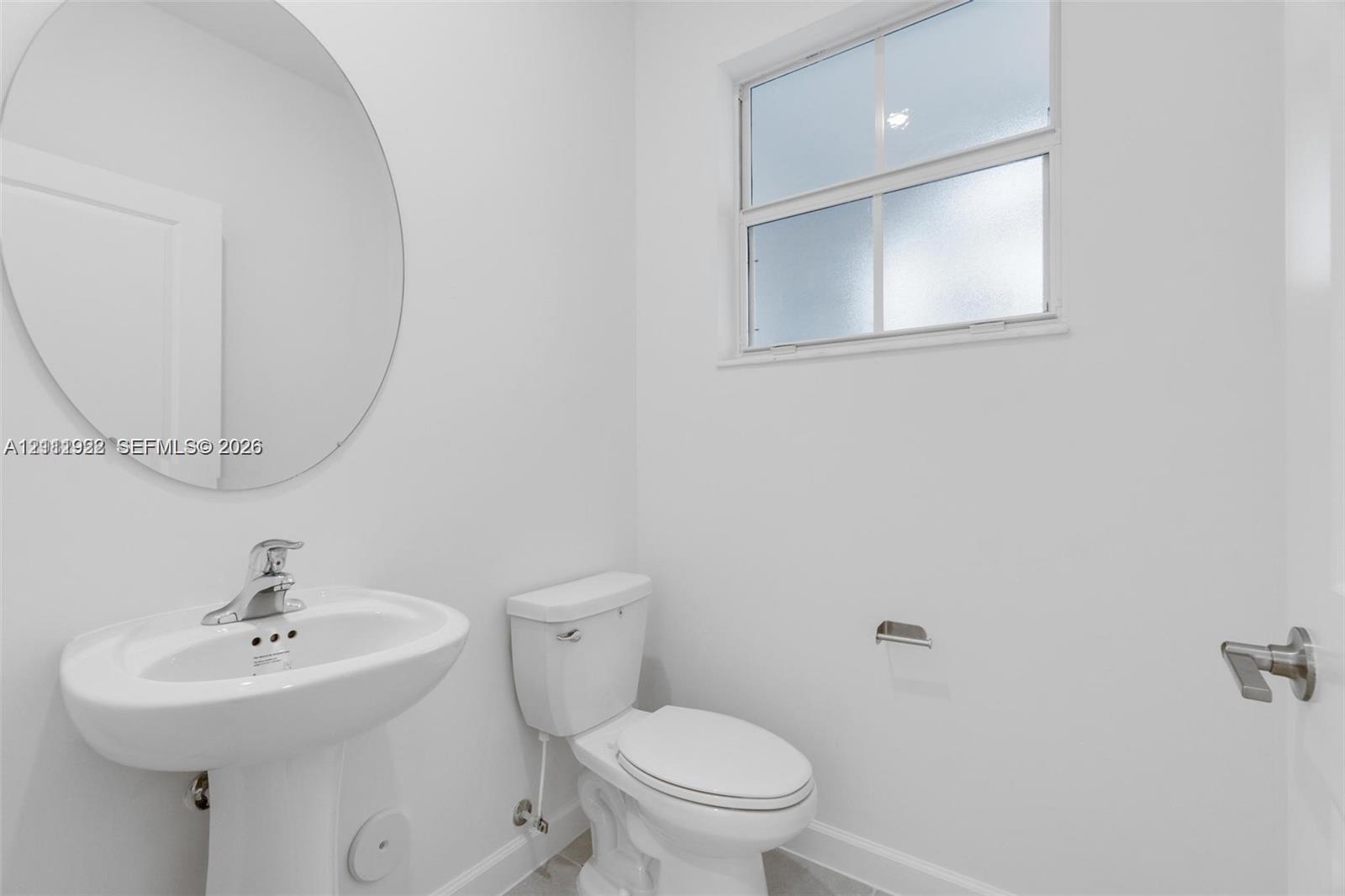 Pedestal sink featuring a polished chrome faucet, round wall-mounted mirror, dual-flush toilet, and natural light via an obscured glass window