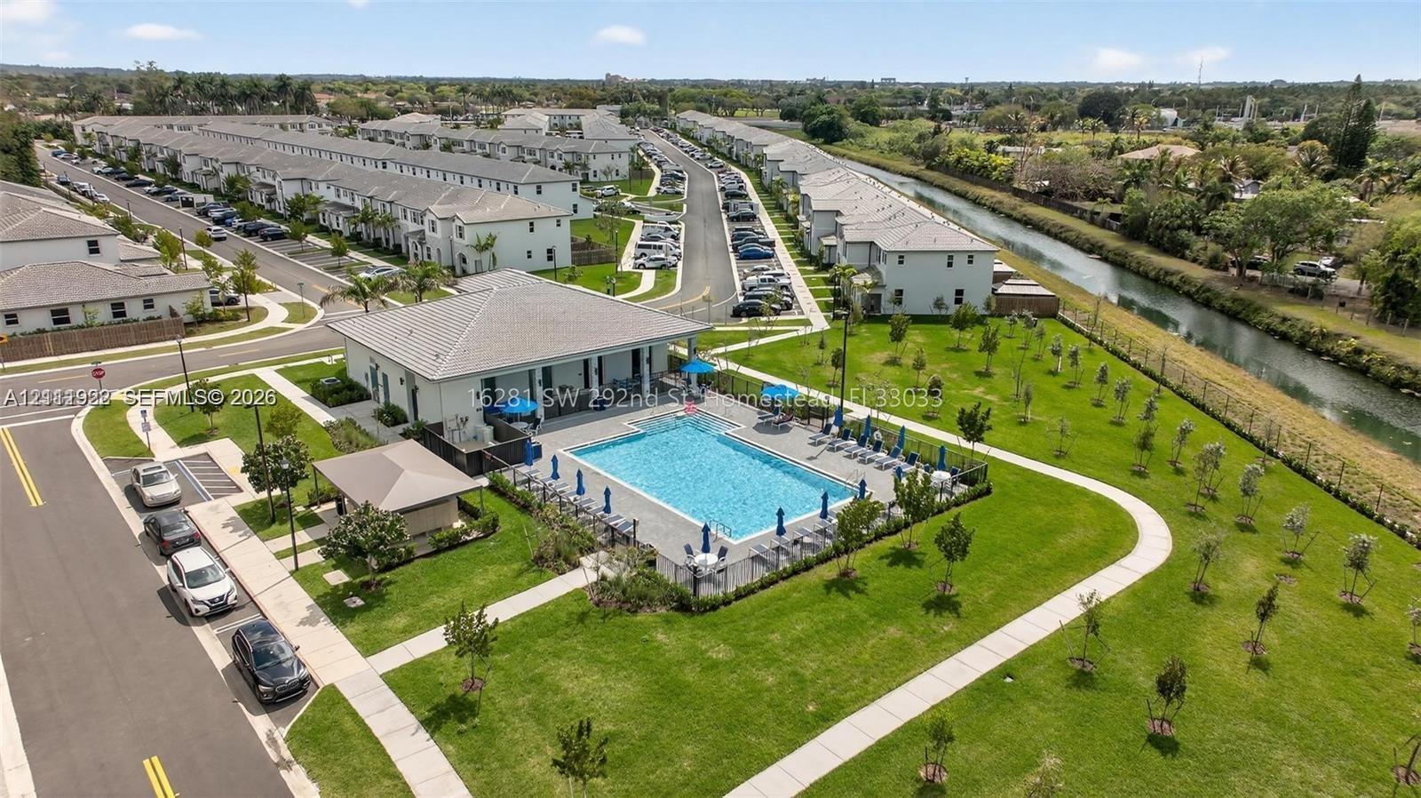 Community swimming pool with a surrounding paved deck, covered pavilion structure, manicured green space, and concrete walking paths
